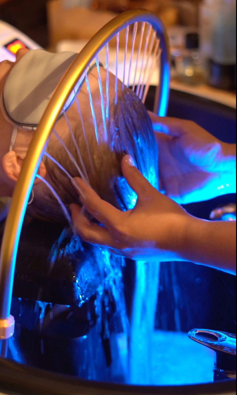 A person lying down with their head in a spa basin while another person washes their hair under blue LED lighting. A gold-framed hair rinsing device is positioned over the head.