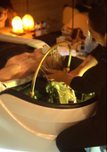 A person receives a facial spray massage in a spa setting, leaning back in a white treatment basin with a gold faucet while another person applies pressure. Warm glowing salt lamps and dim ambient lighting create a relaxing atmosphere.